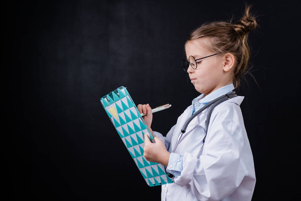 Adorable little girl in whitecoat making prescriptions for her patients in medical document in front of camera - Fotó, kép