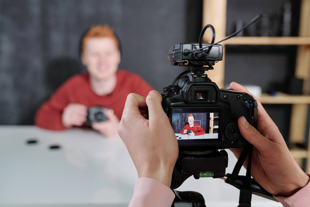 Manos del operador de video sosteniendo la cámara frente al blogger masculino mostrando nuevo equipo fotográfico mientras está sentado junto al escritorio
 - Foto, imagen