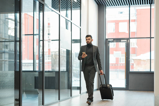 Young elegant businessman scrolling in smartphone while moving along airport and pulling suitcase with luggage - Foto, imagen