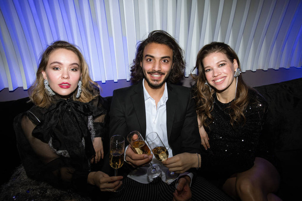 Three young glamorous friends with flutes of champagne sitting on couch in night club, toasting and enjoying party - Valokuva, kuva