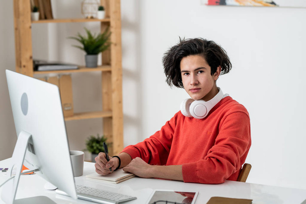 Young casual male freelancer looking at you while making notes by desk in front of computer screen at home - Photo, Image