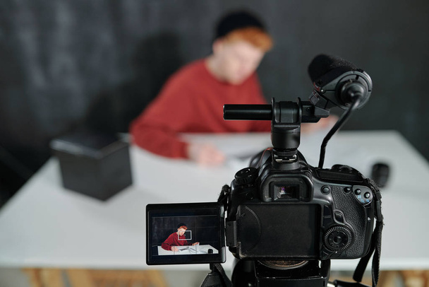 Bildschirm der Videokamera vor dem jungen männlichen Vlogger, der am Schreibtisch vor schwarzem Hintergrund im Studio sitzt - Foto, Bild