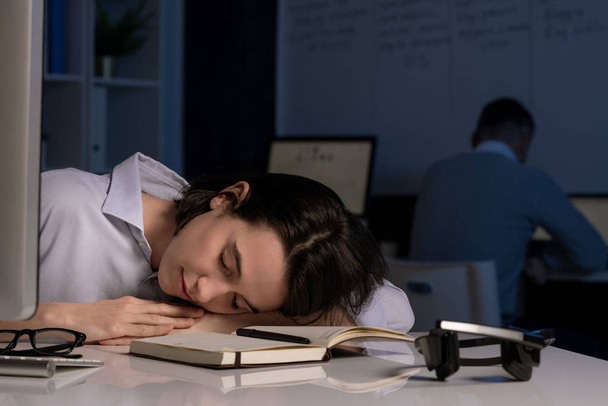 Agotada joven operadora de línea directa durmiendo con la cabeza en el escritorio después de trabajar hasta tarde en la noche con su colega de fondo
 - Foto, Imagen