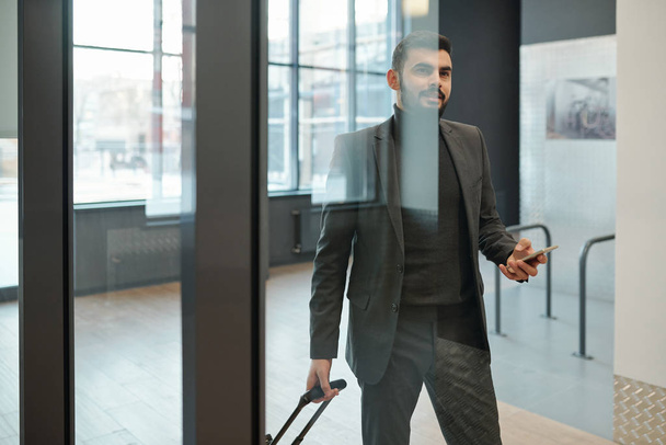 Giovane uomo d'affari contemporaneo in abbigliamento formale con valigia in piedi a porta in aeroporto durante il viaggio
 - Foto, immagini