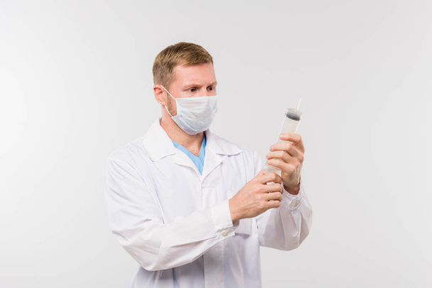 Jonge mannelijke clinicus in witte jas en beschermend masker met grote spuit voor de medische procedure - Foto, afbeelding