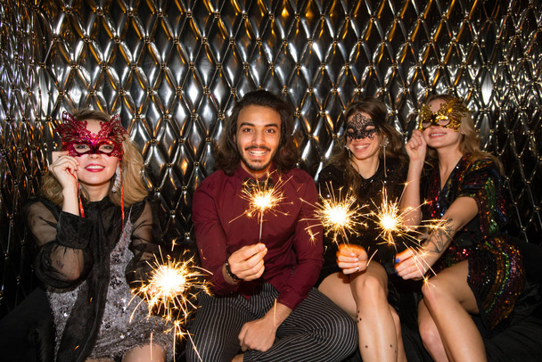 Three happy girls in venetian masks and guy holding sparkling bengal lights while sitting by wall in the night club - Photo, Image