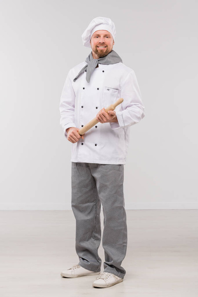 Felice giovane chef barbuto in uniforme che tiene il mattarello di legno mentre si trova di fronte alla telecamera in isolamento
 - Foto, immagini