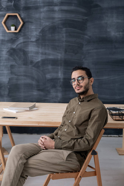 Młody poważny biznesmen w ubraniach casualwear i okularach siedzi na krześle przy stole na tle tablicy - Zdjęcie, obraz