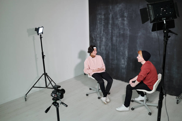 Two happy young vloggers in casualwear sitting on chairs in front of each other against black background in studio - Photo, Image