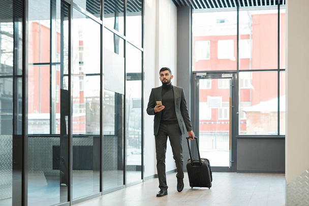 Young elegant businessman with smartphone pulling suitcase with luggage while moving along airport - Fotoğraf, Görsel