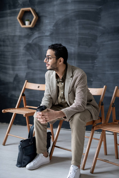 Mladý podnikatel se sklenicí kávy a kabelky sedí na židli v kavárně nebo kanceláři na pozadí tabule - Fotografie, Obrázek