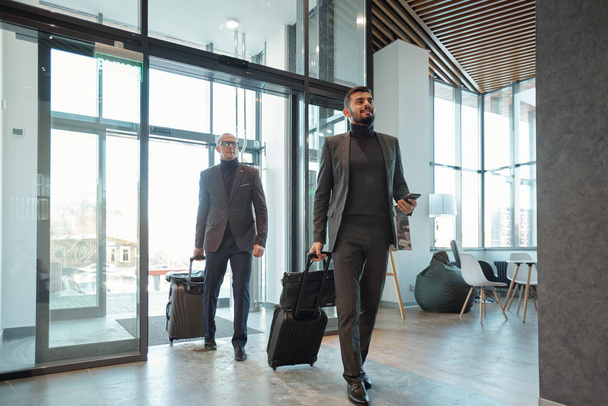 Due eleganti viaggiatori d'affari tirando valigie mentre entrano nella lounge dell'hotel dopo l'arrivo in paese straniero
 - Foto, immagini