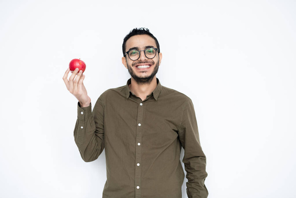 Happy young man with toothy smile holding ripe red apple in right hand while standing in front of camera - Photo, Image