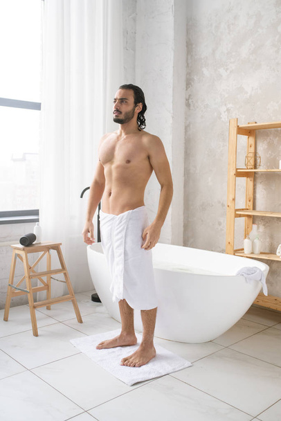 Young shirtless man with soft white towel on hips standing on small rug after having hot bath with foam - Photo, image