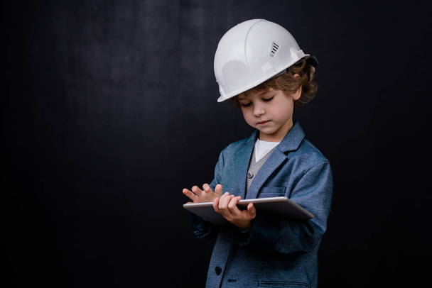 Serious cute little boy in hardhat and formalwear using digital tablet while surfing in the net against black background - Fotografie, Obrázek