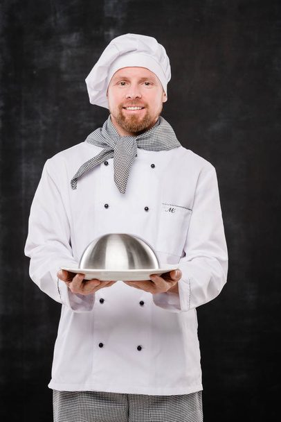 Młody uśmiechnięty kucharz w mundurze trzymający klosz z gotowanym posiłkiem dla Ciebie stojąc w izolacji na czarnym tle - Zdjęcie, obraz