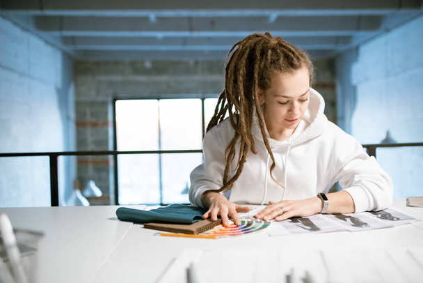 Młoda projektantka mody patrząc na szkice na biurku przy wyborze kolorów do nowej kolekcji w studio - Zdjęcie, obraz