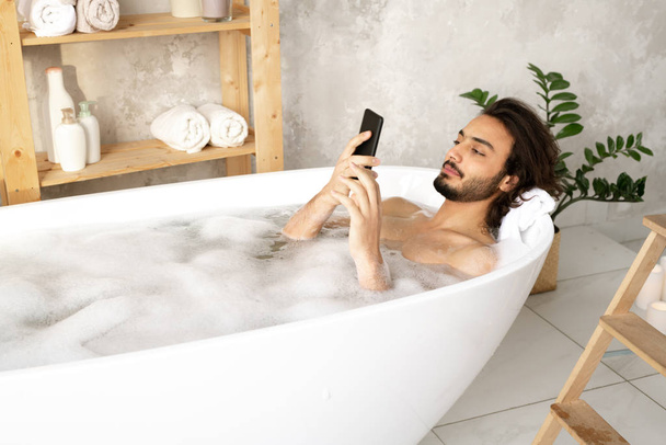 Young naked man watching video or scrolling in smartphone while lying in bathtub filled with water and foam in bathroom - Fotó, kép