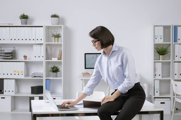 Genç zarif iş kadını, masa başında oturuyor ve internette bilgi ararken dizüstü bilgisayarına hafifçe eğiliyor. - Fotoğraf, Görsel