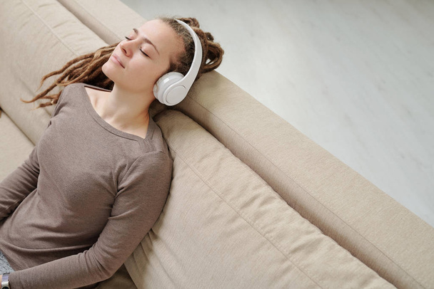 Cute kobieta ze słuchawkami korzystających muzyki na relaks siedząc na wygodnej kanapie po dniu roboczym - Zdjęcie, obraz