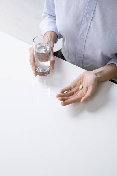 Egy beteg, inges nő vagy üzletasszony kezei, akik egy pohár vizet és pirulát fognak inni, miközben az asztalnál ülnek. - Fotó, kép