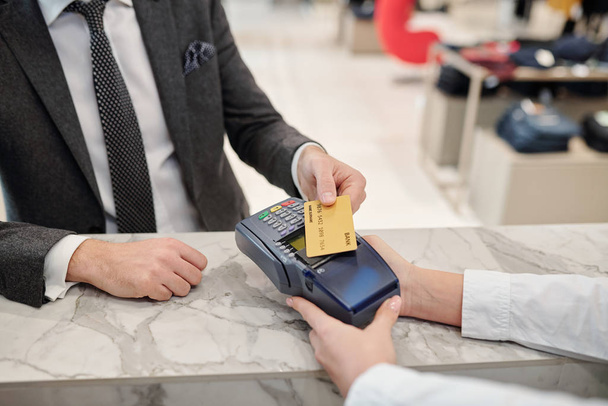 Handen van de jonge elegante man met een creditcard over de betaalautomaat terwijl het kopen van iets in de hedendaagse boetiek - Foto, afbeelding