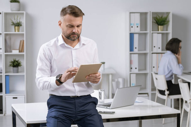 Trabalhador de escritório sério ou empregador em formalwear usando touchpad enquanto navega na rede para obter informações necessárias
 - Foto, Imagem