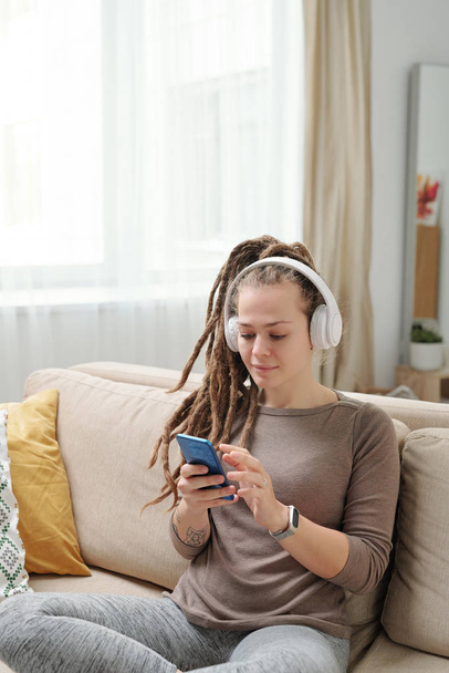 Young female with dreadlocks scrolling in smartphone while sitting on couch and relaxing at leisure - Φωτογραφία, εικόνα