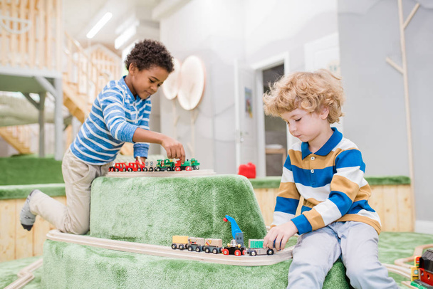 Dos niños pequeños interculturales sentados en el patio de recreo mientras juegan trenes de juguete juntos en el centro infantil contemporáneo
 - Foto, imagen