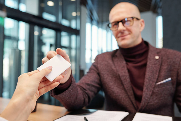 Hand van kale, volwassen zakenman in brillen en formele kleding die een kaart neemt uit een lege hotelkamer bij de receptie - Foto, afbeelding