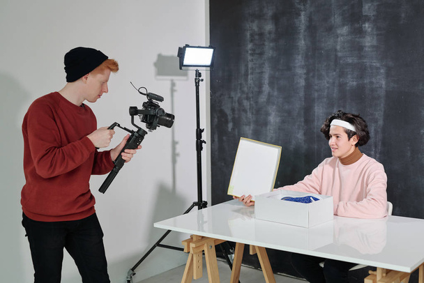 Vakava nuori mies, jolla on videokuvauslaitteet seisomassa ystävänsä edessä, istuu pöydän ääressä ja avaa laatikon uusilla jalkineilla
 - Valokuva, kuva
