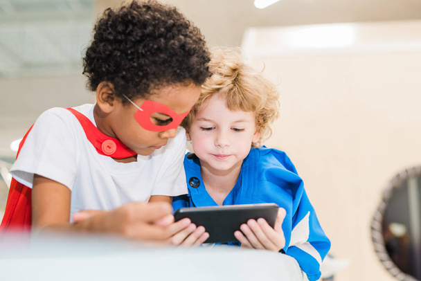 Twee kleine interculturele vriendelijke jongens in kostuums van superman kijken nieuwsgierige video in smartphone na het spelen - Foto, afbeelding