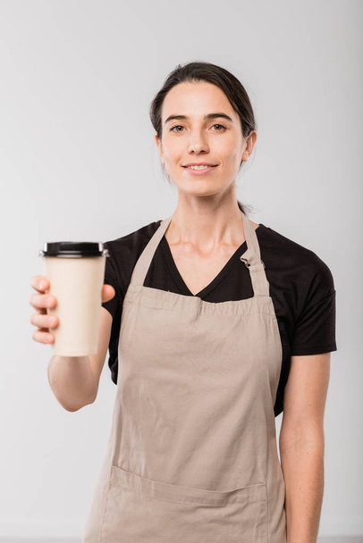 Jonge glimlachende serveerster van cafe geven u papier glas hete koffie terwijl staan voor de camera in isolatie - Foto, afbeelding