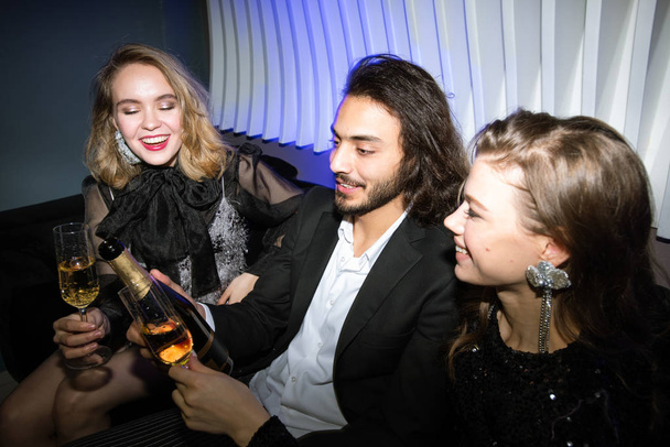 Gelukkig glamoureuze meisjes met fluiten van champagne en jonge man houden fles terwijl zitten op de bank in de nachtclub en genieten van feest - Foto, afbeelding