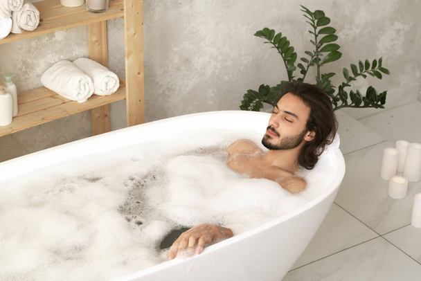 Young relaxed man with closed eyes lying in white bathtub filled with hot water and foam with wooden shelves near by - Photo, Image