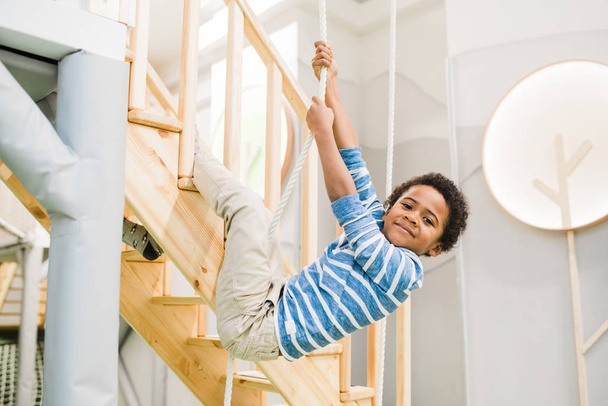 Happy little cute boy of African ethnicity holding by rope while hanging over wooden staircase during play - Foto, afbeelding