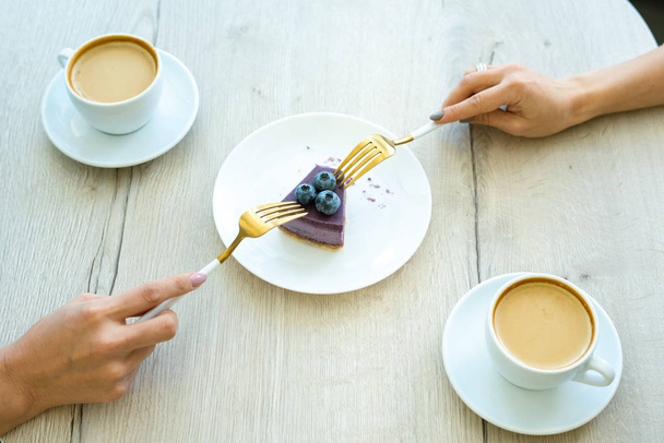 Χέρια των δύο κοριτσιών με πιρούνια λαμβάνοντας κομμάτι νόστιμο cheesecake βατόμουρο στο πιάτο, ενώ έχοντας καπουτσίνο στο καφέ - Φωτογραφία, εικόνα
