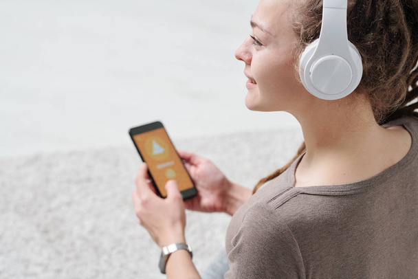 Mulher jovem bonito em fones de ouvido e activewear segurando smartphone enquanto ouve aulas de áudio da prática de ioga
 - Foto, Imagem