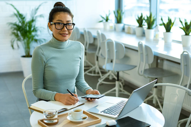 Jonge succesvolle zakenvrouw in casualwear zitten aan tafel in cafe, werken voor de laptop en het maken van notities - Foto, afbeelding