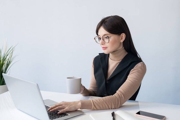 Młoda, poważna bizneswoman z kubkiem analizującym online dane finansowe siedząc przed laptopem w biurze - Zdjęcie, obraz