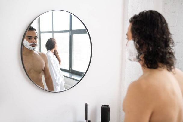 hemdloser junger Mann mit Rasierschaum am Bart, der vor dem Spiegel steht und seine Freundin im weißen Bademantel reflektiert - Foto, Bild