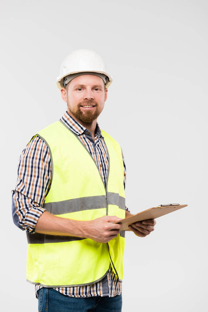 Cheerful young foreman or engineer with clipboard looking at you while standing in front of camera - Photo, Image