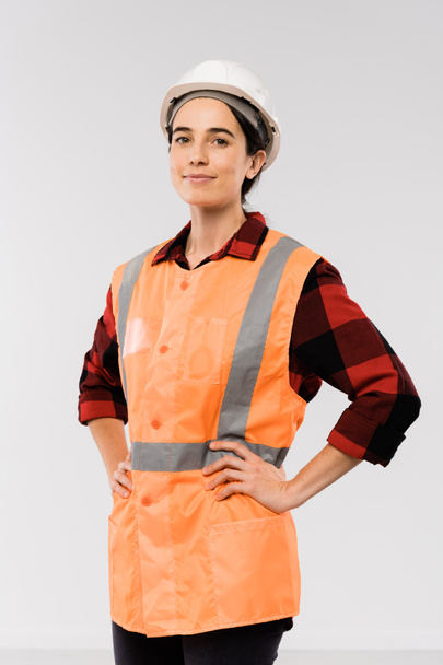 Bella giovane ingegnere donna in casco e giacca arancione tenere le braccia in vita mentre in piedi di fronte alla fotocamera
 - Foto, immagini