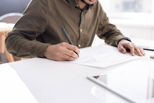 Νεαρός απασχολημένος οικονομολόγος σχέδιο διάγραμμα ροής στο χαρτί, ενώ κάθεται από το γραφείο στο γραφείο και την προετοιμασία της έκθεσης - Φωτογραφία, εικόνα