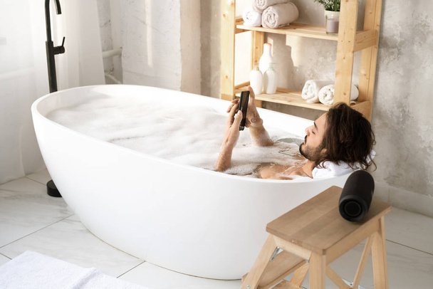Молодой спокойный мужчина с помощью смартфона, лежа в ванне, наполненной водой и пеной в углу ванной комнаты
 - Фото, изображение