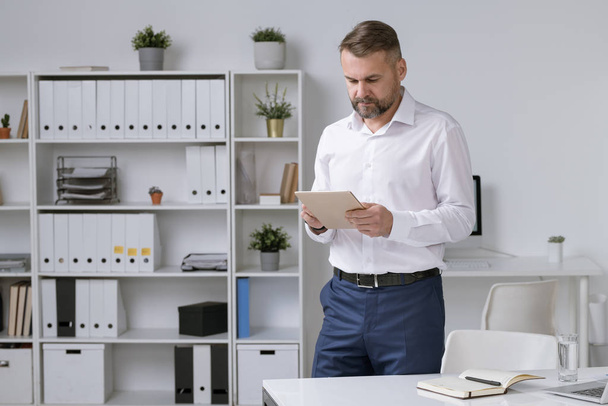 Empreendedor maduro confiante rolando em tablet digital enquanto procura por dados on-line ou assistindo webinar no escritório
 - Foto, Imagem