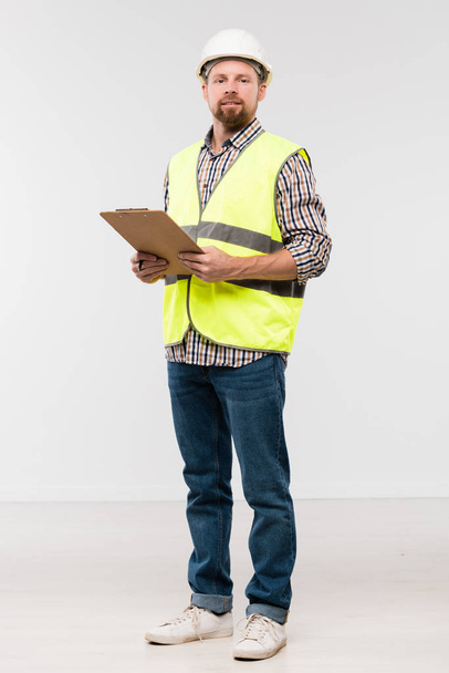 Bearded young engineer with clipboard standing in front of camera in isolation and looking at you - Foto, Imagen
