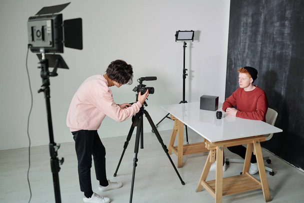 Genç kameraman, erkek vlogger 'ın önünde stüdyoda dikilirken video çekim ekipmanlarının önünde eğiliyor. - Fotoğraf, Görsel