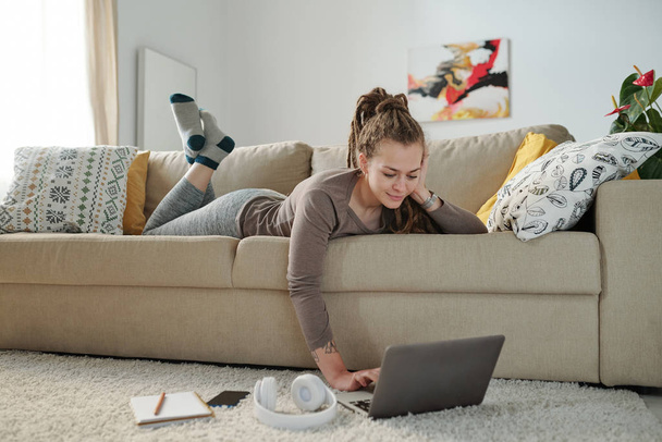 Счастливая спокойная девочка лежит на диване и смотрит на дисплей ноутбука на полу, оставаясь дома в выходные
 - Фото, изображение