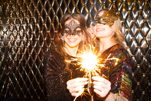 Dwie wesołe dziewczyny w weneckich maskach trzymające błyszczące bengalskie światła podczas zabawy w klubie nocnym - Zdjęcie, obraz